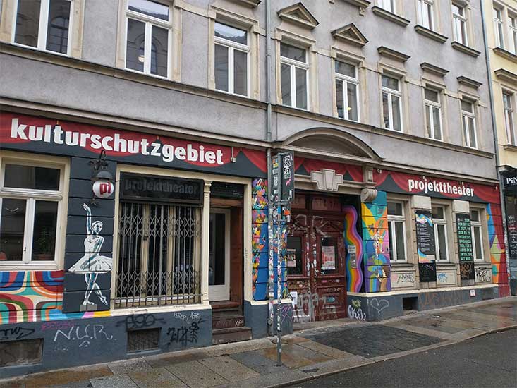 Außenansicht des Eingangs des Projekttheaters auf der Louisenstraße: historisches Gebäude mit bunter Fassade, Schaufenstern und davor Gehweg.