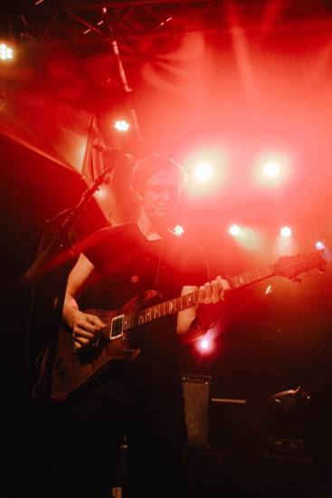 Person spielt E-Gitarre auf einer Bühne, umgeben von rotem Licht und Scheinwerfern. Die Szene wirkt energiegeladen und atmosphärisch.