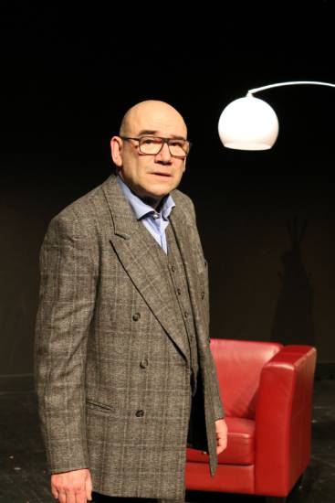 Ein Mann mit Brille im grauen Anzug und blauen Hemd steht auf einer Theaterbühne. Hinter ihm befinden sich ein roter Sessel und eine weiße Stehlampe.