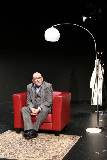 Ein Mann mit Brille im grauen Anzug sitzt auf einem roten Sessel auf der Theaterbühne. Neben ihm steht eine weiße Bogenlampe, rechts im Hintergrund hängt ein heller Mantel an einer Garderobe.