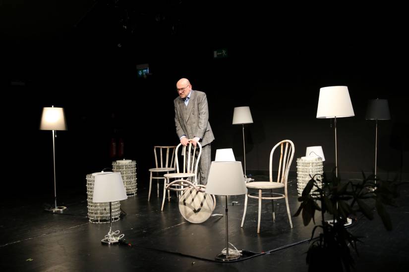 Ein Mann im grauen Anzug steht auf einer Theaterbühne und stützt sich auf eine Holzstuhllehne. Um ihn herum stehen mehrere Stühle und verschiedene Lampen, die eine wohnzimmerähnliche Atmosphäre erzeugen.