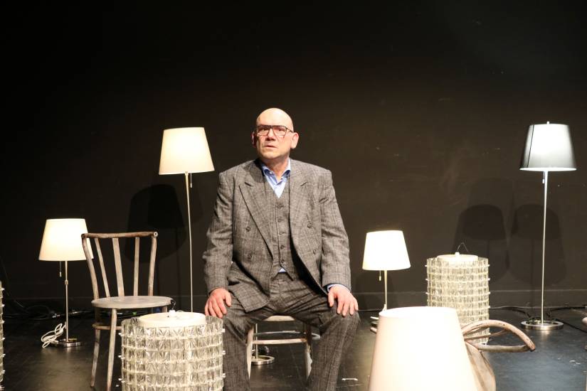 Ein Mann im grauen Anzug sitzt auf einem Stuhl auf einer Theaterbühne. Um ihn herum stehen mehrere Lampen und Stühle, die als Bühnenbild arrangiert sind.