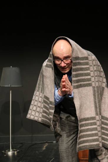Ein Mann mit Brille im grauen Anzug steht auf einer Theaterbühne. Er hat sich eine große grau karierte Decke über den Kopf gelegt und hält die Hände vor der Brust in einer Geste des Dankes oder Gebets. Im Hintergrund ist eine Stehlampe zu sehen.