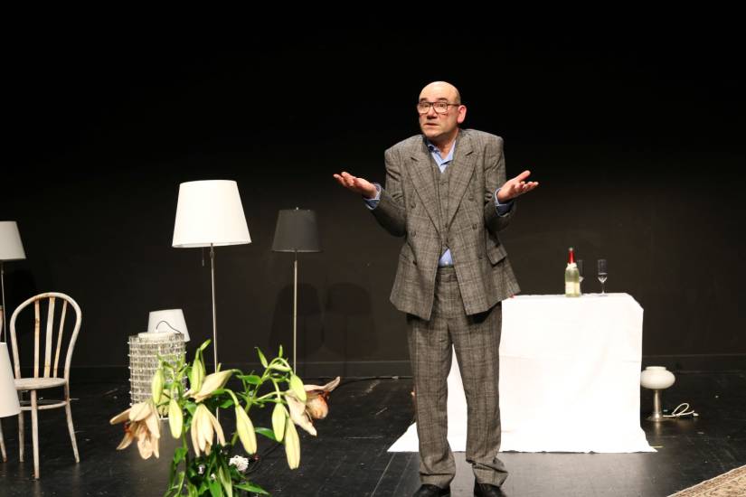 Ein Mann im grauen Anzug mit Brille steht auf einer Theaterbühne. Er hebt die Arme fragend zur Seite. Um ihn herum stehen Stühle, Stehlampen, ein Tisch mit weißer Tischdecke, einer Flasche Wein und einem Glas. Im Vordergrund sind Lilienblüten zu sehen.