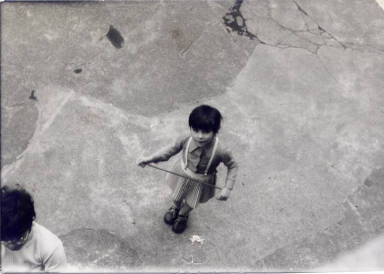 Schwarz-weiß-Fotografie eines Kindes, das von oben aufgenommen wurde. Das Kind hält ein Springseil in den Händen und schaut nach oben in die Kamera. Links unten ist der Kopf einer weiteren Person angeschnitten.