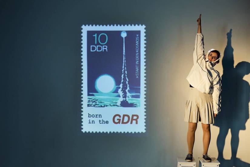 Person in weißem Kostüm mit Faltenrock und Kopfbedeckung steht auf einem Hocker und streckt den Arm nach oben. Links daneben ist eine große Projektion einer DDR-Briefmarke mit Raketenmotiv und der Aufschrift ‚10 DDR – born in the GDR‘ zu sehen.