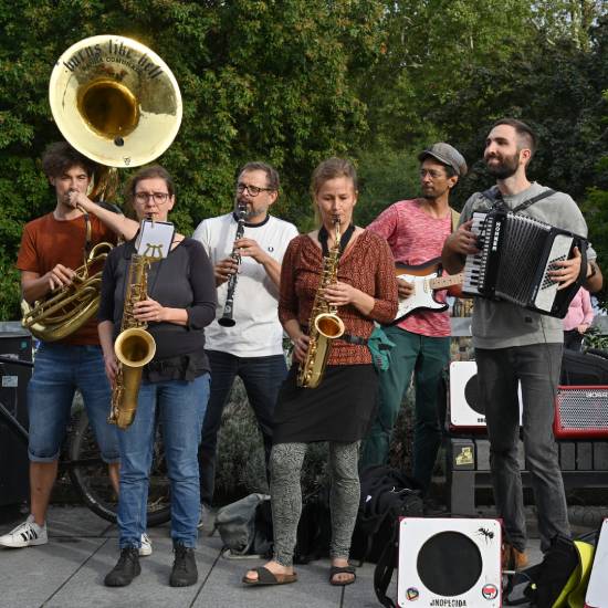 Straßenmusikgruppe mit sieben Musikerinnen und Musikern, die verschiedene Instrumente spielen, darunter Tuba, Saxophone, Klarinette, E-Gitarre und Akkordeon. Sie treten im Freien vor Bäumen auf.