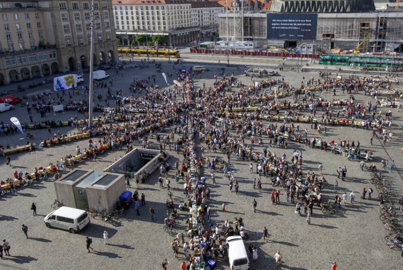 Große Menschenmenge auf dem Altmarkt in Dresden, die an langen, sternförmig angeordneten Tafeln zusammensitzt. Im Hintergrund sind historische Gebäude, Straßenbahnen und eine Baustelle zu sehen.