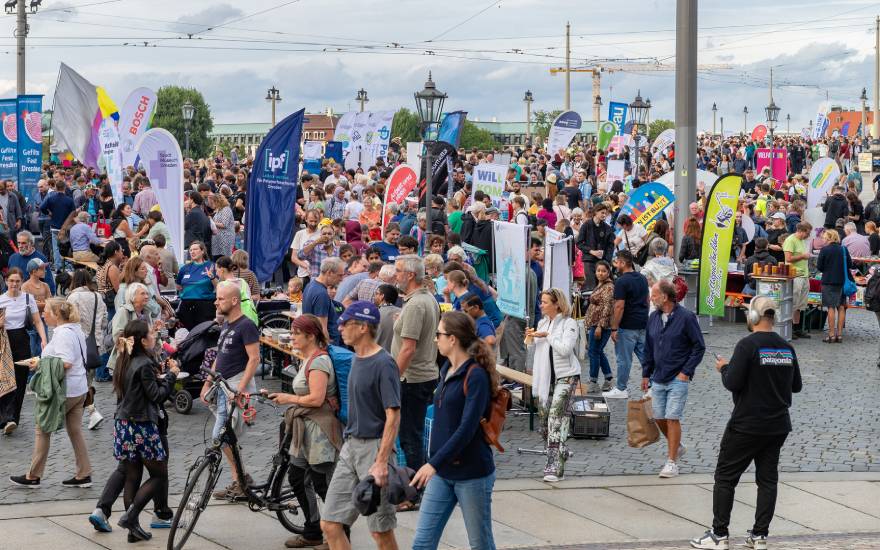 Menschenmenge auf einem Platz in Dresden bei einem bunten Stadtfest. Zahlreiche Infostände und Banner verschiedener Organisationen sind aufgebaut, Besucher schlendern, essen und unterhalten sich.