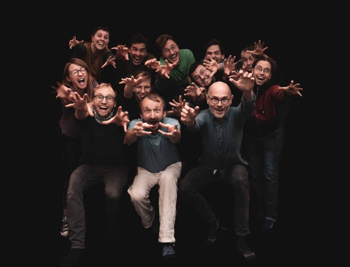 Gruppenfoto von dreizehn Personen vor schwarzem Hintergrund, die lachend mit ausgebreiteten Armen und gespreizten Fingern in die Kamera greifen, als wollten sie das Publikum erschrecken oder umarmen.