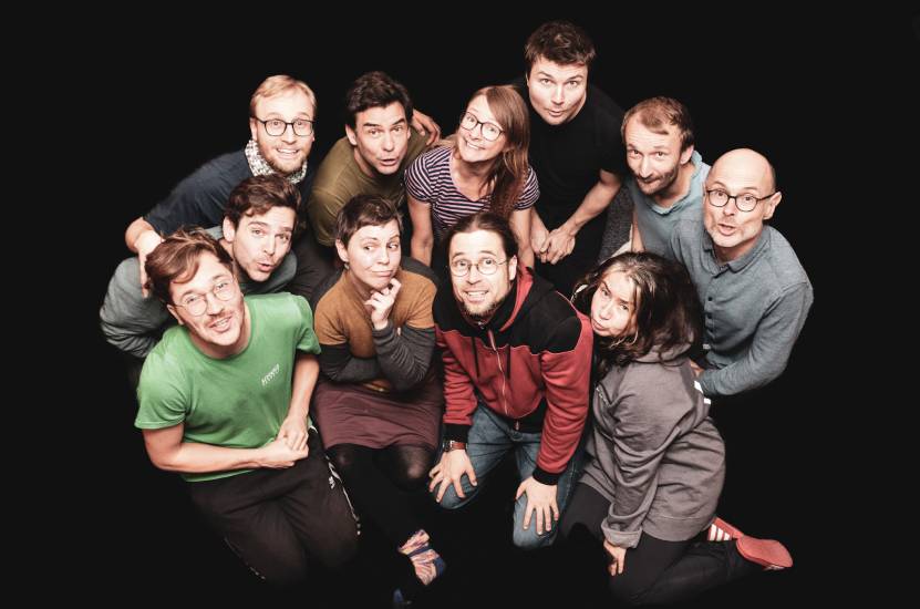 Gruppenfoto von elf Personen, die eng zusammengerückt und von oben fotografiert sind. Alle schauen lachend oder mit witzigen Gesichtern in die Kamera, vor schwarzem Hintergrund.