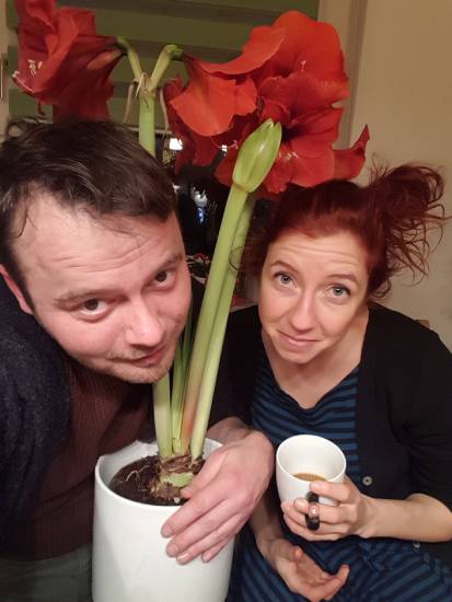 Ein Mann und eine Frau sitzen nebeneinander in einer Wohnung. Vor ihnen steht ein Blumentopf mit großen roten Blüten. Der Mann lehnt sich leicht nach vorn, die Frau hält eine weiße Tasse in der Hand und schaut in die Kamera.