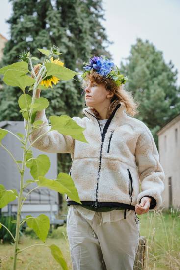 Person mit Blumenkranz auf dem Kopf und heller Fleecejacke betrachtet eine Sonnenblume und berührt deren Blätter.