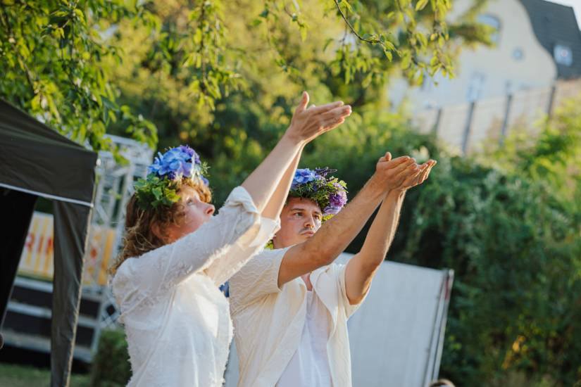 Zwei Personen mit Blumenkränzen auf dem Kopf heben im Freien die Arme nach oben, als ob sie etwas auffangen oder begrüßen.
