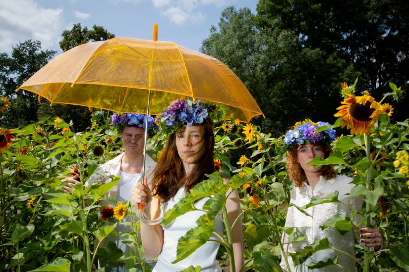 Drei Personen stehen in einem Sonnenblumenfeld. Sie tragen weiße Kleidung und Blumenkränze auf dem Kopf. Eine Person in der Mitte hält einen großen, durchsichtigen gelben Regenschirm.