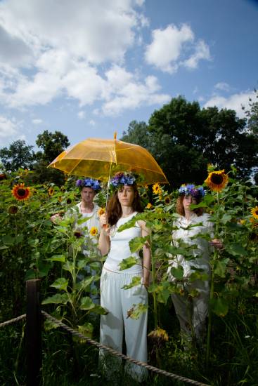 Drei Personen stehen in einem Sonnenblumenfeld. Sie tragen weiße Kleidung und Blumenkränze auf dem Kopf. Die Person in der Mitte hält einen großen gelben Regenschirm über sich.