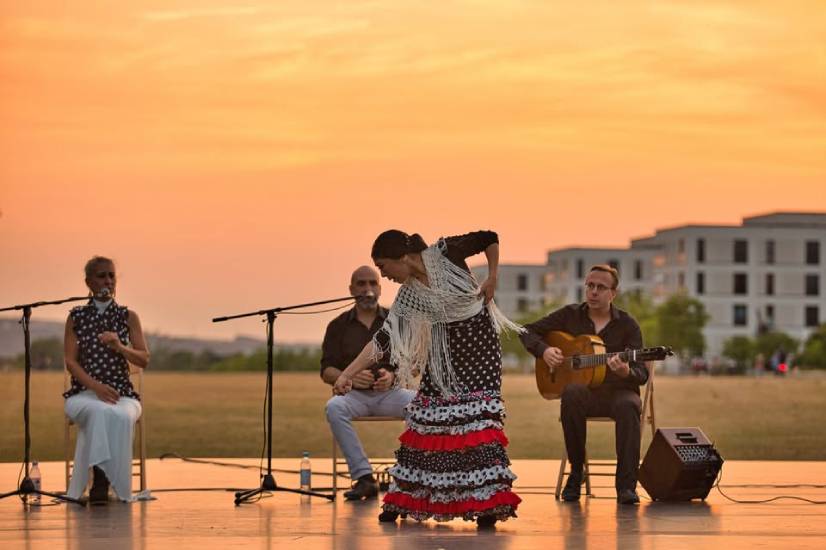 Flamenco-Aufführung im Freien bei Sonnenuntergang: Eine Tänzerin im schwarzen Kleid mit weißen Punkten, roten Rüschen und weißem Fransenschal tanzt vor drei Musikern mit Mikrofonen und Gitarre. Im Hintergrund moderne Gebäude und ein orange leuchtender Himmel