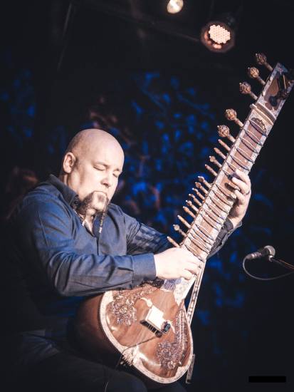 Mann in dunklem Hemd spielt auf einer großen Sitar auf einer Bühne. Er ist konzentriert, die Bühne ist in blaues Licht getaucht.