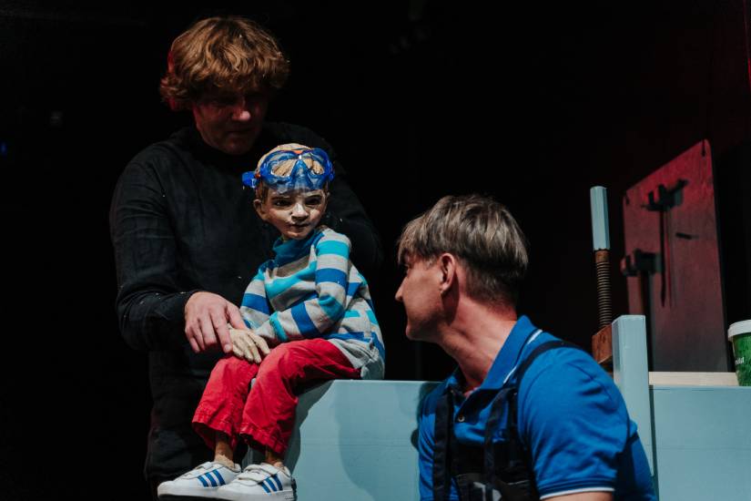 Eine Kinderpuppe mit Taucherbrille, gestreiftem Pullover und roter Hose sitzt auf einem blauen Podest. Hinter ihr steht ein Puppenspieler in Schwarz, der sie führt. Ein weiterer Darsteller in blauer Kleidung kniet davor und blickt die Puppe freundlich an.