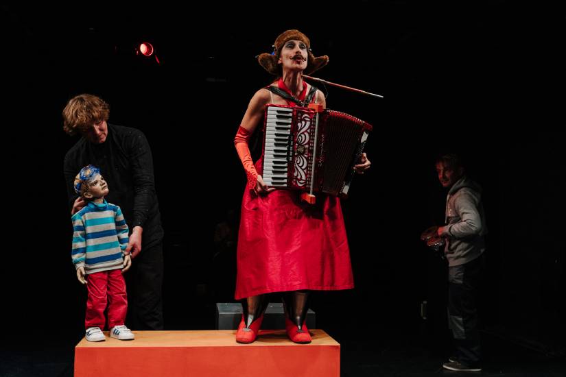 Auf einer Theaterbühne steht eine Frau in einem roten Kleid mit Akkordeon und Zigarettenspitze auf einem Podest. Links daneben führt ein Puppenspieler eine Kinderpuppe im gestreiften Pullover und roten Hosen. Im Hintergrund rechts steht ein weiterer Darsteller mit Requisiten.