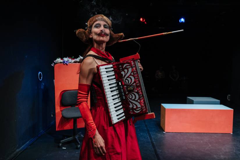 Eine Person in rotem Kleid und langen roten Handschuhen steht auf einer Theaterbühne, trägt ein Akkordeon vor dem Körper und einen Zigarettenhalter im Mund. Sie ist stark geschminkt und blickt direkt ins Publikum.