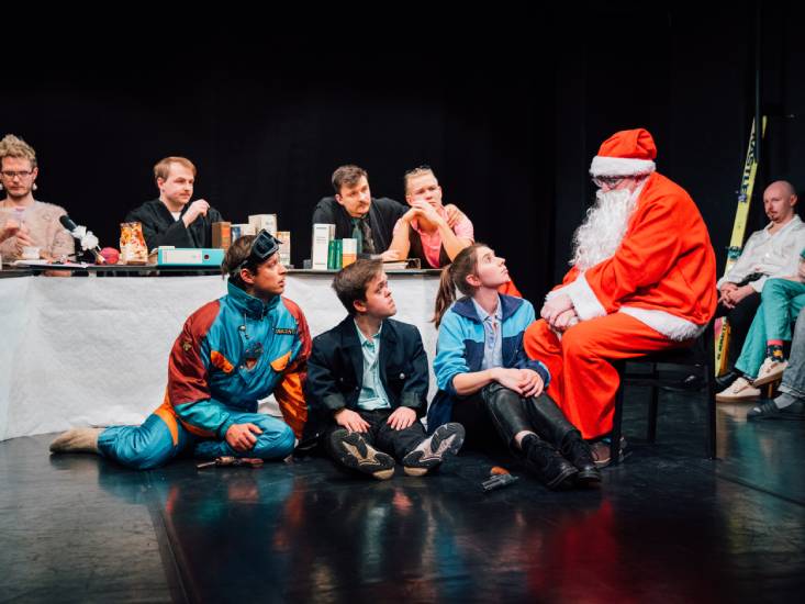 neun Personen sitzen auf einer Bühne verteilt. Vier Person sitzen an einem Tisch. Eine Person sitzt im Hintergrund. Drei Personen sitzen auf dem Boden und schauen zu einer weiteren Person auf, die im Weihnachtsmann-Kostüm auf einem Hocker daneben sitzt.