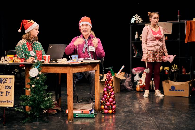 3 Personen auf einer weihnachtlich geschmückten Bühne. Eine Person steht im Hintergrund und hält ein Kuscheltier und eine Klobürste in den Händen. Zwei Personen sitzen im Vordergrund an einem Tisch. Die Person rechts spricht. Die linke Person hört zu und sieht sie an.