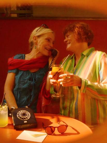 Zwei Frauen sitzen an einem runden orangefarbenen Tisch in entspannter Atmosphäre. Die Frau links trägt eine blaue Bluse, eine rote Stola und hält einen alten Telefonhörer ans Ohr. Die Frau rechts trägt ein bunt gestreiftes Hemd und hält ein Glas mit Orangenscheibe in der Hand. Auf dem Tisch stehen Getränke, eine Sonnenbrille und ein schwarzes Wählscheibentelefon. Beide lachen sich herzlich an.