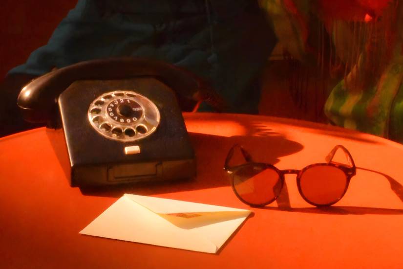 Nahaufnahme eines orangefarbenen Tisches mit einem schwarzen Wählscheibentelefon, einer braunen Sonnenbrille und einem weißen Briefumschlag. Die Szene wirkt durch warmes Licht nostalgisch und retro.