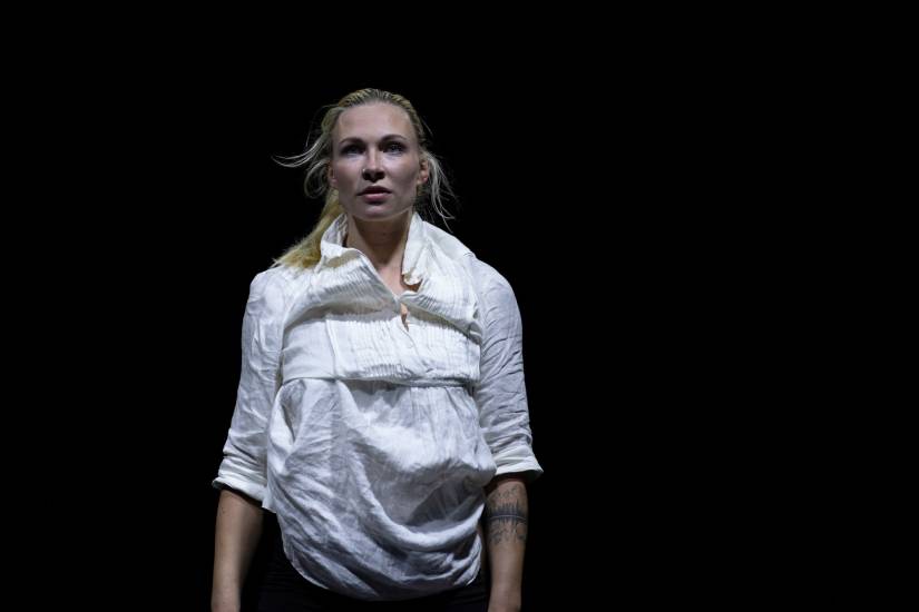 Person mit blondem Pferdeschwanz steht im schwarzen Bühnenraum. Sie trägt ein weißes, locker sitzendes Hemd mit Faltenwurf und blickt ernst nach vorn.