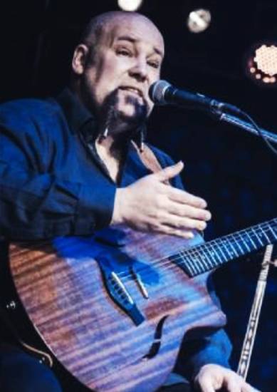 Mann mit Gitarre sitzt auf einer Bühne und singt in ein Mikrofon. Er trägt ein dunkles Hemd und gestikuliert mit der Hand.