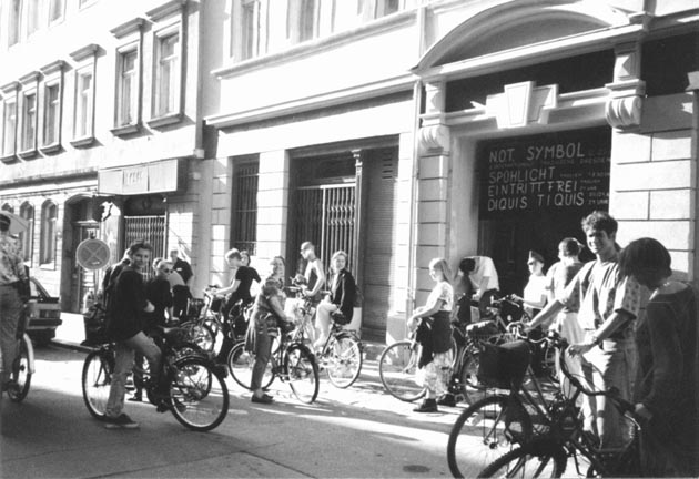 Schwarz-Weiß-Aufnahme der Frontfassade des Projekttheaters Dresden gegen Ende des 20. Jahrhundert. Davor stehen viele junge bis mittelalte Menschen mit Fahrrädern. Einige von ihnen schauen direkt in die Kamera.