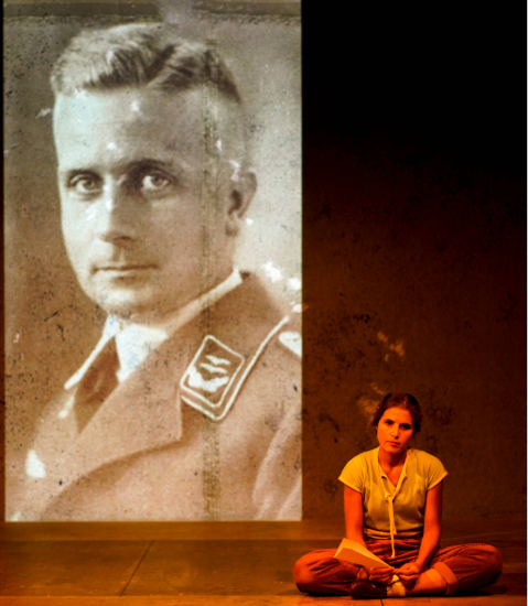Eine Frau sitzt vor der Projektion eines historischen schwarz-weiß Fotos. Sie sitzt mit dem Rücken zum Foto und blickt geradeaus in die Kamera. Das Foto zeigt das Porträt eines Mannes im mittleren Alter. Er trägt eine Uniform.