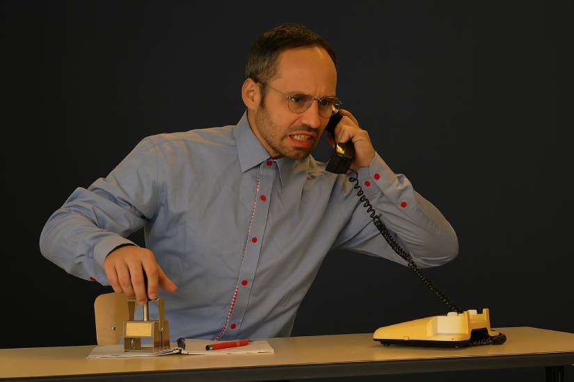 Ein Mann in grauem Hemd sitzt an einem Tisch. Er spricht aufgebracht in ein altes Telefon. Das Telefon steht vor ihm auf dem Tisch. Den Hörer hält er in der linken Hand. Mit der rechten Hand drückt er einen Stempel auf die Seite eines Ringblocks.
