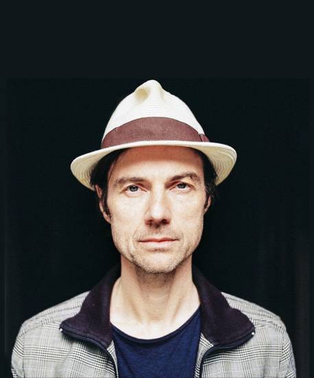 Porträtfoto des Musikers Jörg Holdinghausen vor einem schwarzen Hintergrund. Jörg Holdinghausen trägt Hut und schaut direkt in die Kamera.