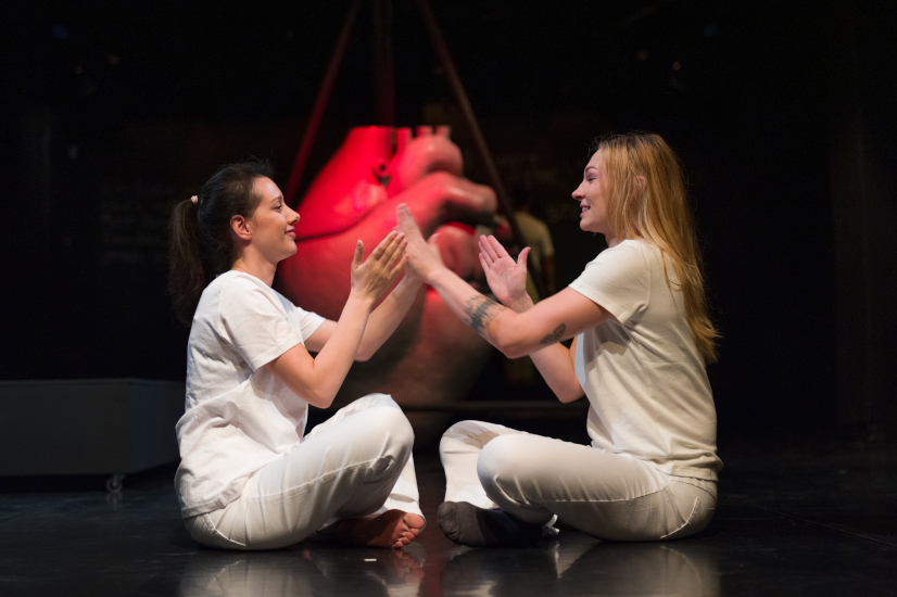 Zwei Schauspielerinnen sitzen auf einer Bühne. Sie tragen weiße Kleidung. Sie sitzen im Schneidersitz und schauen sich an. Sie spielen ein Klatschspiel mit den Händen. Beide lachen. Im Hintergrund steht ein großes Herz. Das Herz leuchtet rot.