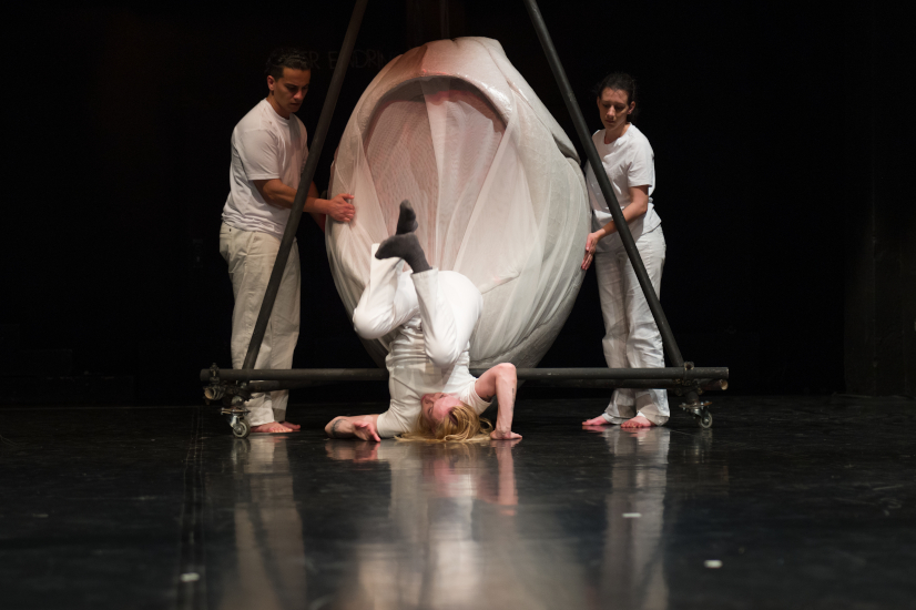 Zwei Schauspieler stützen von zwei Seiten ein großes Herz und blicken auf eine dritte Schauspielerin. Die dritte Schauspielerin ist gerade aus dem Herz gefallen und macht eine Rolle rückwärts.