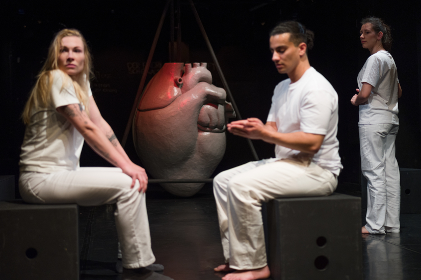 Eine Schauspielerin und ein Schauspieler sitzen sich gegenüber auf schwarzen Würfelhockern. Im Hintergrund beobachtet sie eine weitere Schauspielerin. Dahinter steh ein großes rotes Herz als Bühnenbild.