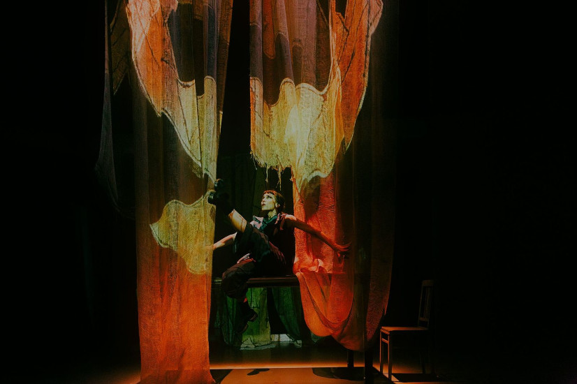 Eine Schauspielerin sitzt auf einer Tischkante und hebt ihr linkes Bein in die Luft. Vor ihr hängt ein transparenter Vorhang auf den große Flügel projiziert werden.