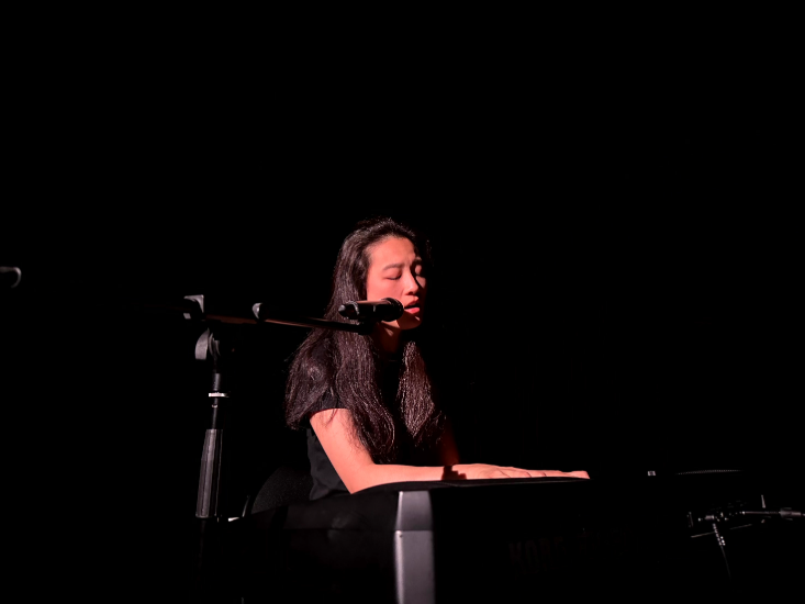 Die Schauspielerin Jing Zhu spielt auf einer dunklen Bühne Klavier und singt in ein Mikrofon.