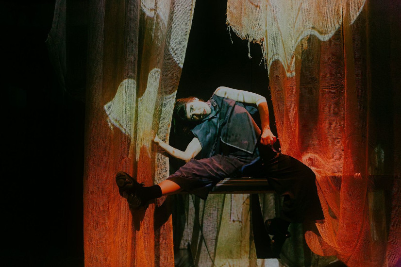 Eine Schauspielerin sitzt mit breiten Beinen auf einer Tischkante und lehnt sich weit über ihr rechtes Bein. Vor ihr hängt ein transparenter Vorhang auf den große Flügel projiziert werden.