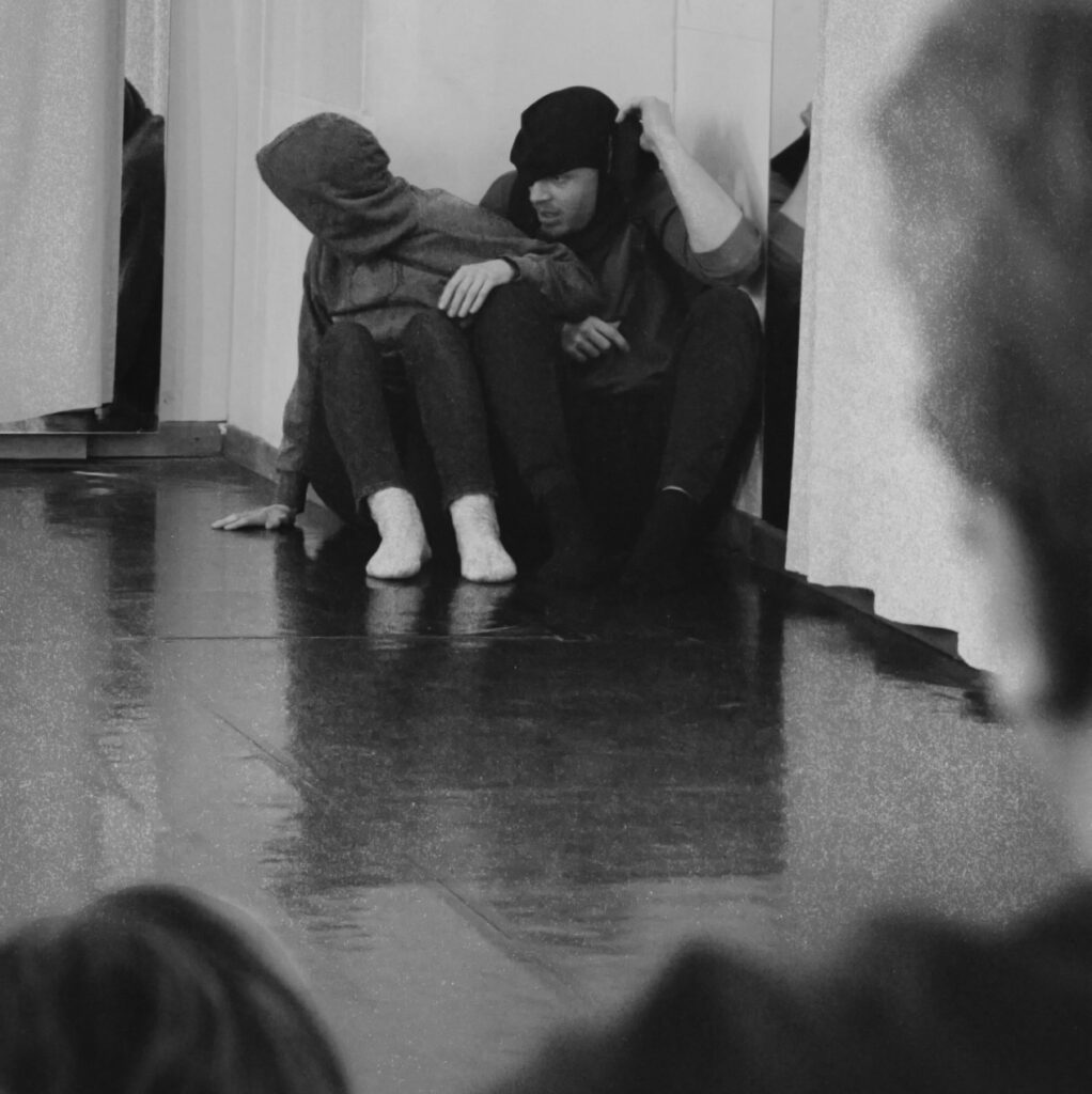 Blick aus dem Publikum auf eine Bühne mit zwei Schauspielern. Sie sind vermummt und sitzen in einer Ecke. Eine Person späht um die Ecke.