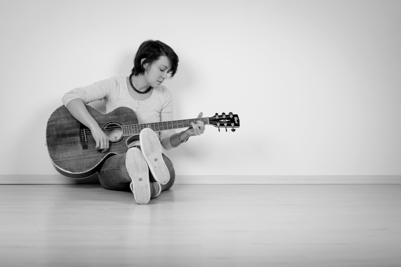 image1550 Hanna Bel sitzt auf dem Boden, lehnt sich an eine weiße Wand und spielt Akustikgitarre