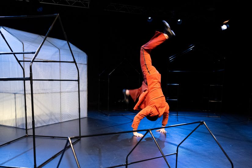 Auf einer schwach blau ausgeleuchteten Bühne macht ein Mann in knallrotem Anzug Handstand.