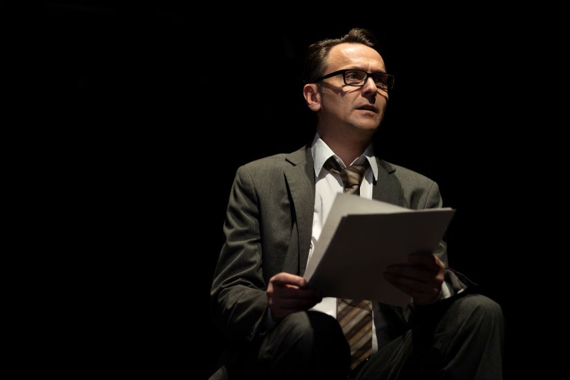 Ein sitzender Mann in Anzug. Er hält Papier in der Hand und schaut nach vorne. Der Hintergrund ist schwarz.