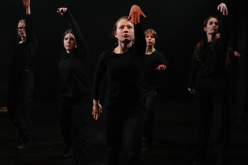 Frontale Aufnahme einer Gruppe junger Schauspielerinnen, die auf einer dunklen Bühne eine Szene performen.