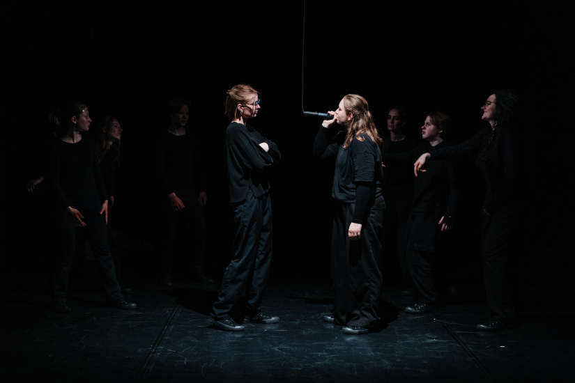 Eine Szene auf einer dunklen Bühne mit einer Gruppe junger Schauspielerinnen. In der Mitte stehen sich zwei Darstellerinnen gegenüber und blicken sich an. Die rechte Schauspielerin rappt in ein Mikrofon.