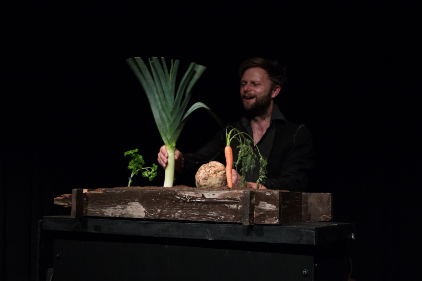 Ein Mann sitzt hinter einem Tisch auf dem ein Kasten mit Erde steht. In der Erde steckt Gemüse. Der Mann bewegt das Gemüse und spielt damit eine Szene in einem Theater für Kinder.
