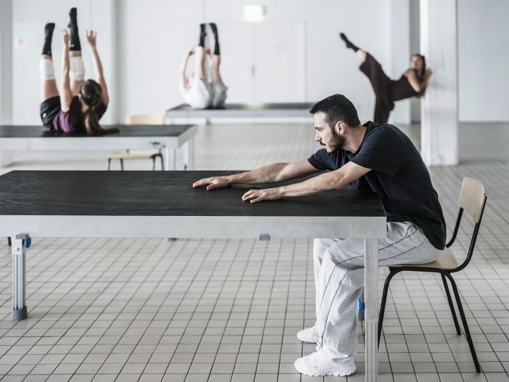 Im Vordergrund sitzt ein Mann an einem Tisch. Er streckt die Arme über die Tischfläche. Im Hintergrund liegen zwei Personen auf Tischen. Sie haben die Arme und Beine in die Luft gestreckt. Eine Frau lehnt an eine Säule. Ein Bein streckt sie in die Luft.