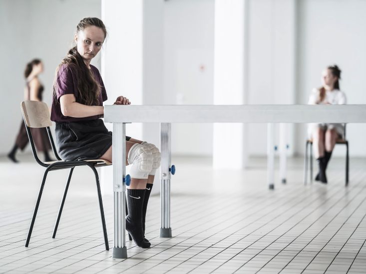 Eine Frau sitzt in einem hellen Raum an einem Tisch. Sie trägt Knieschoner. Im Hintergrund sitzt eine weitere Frau und eine Frau geht vorbei.